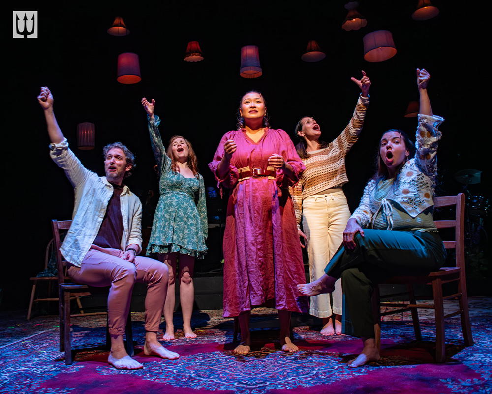 four brightly dressed characters on stage, seated with their arms raised, mouth open wide in song, and Rita standing in the middle