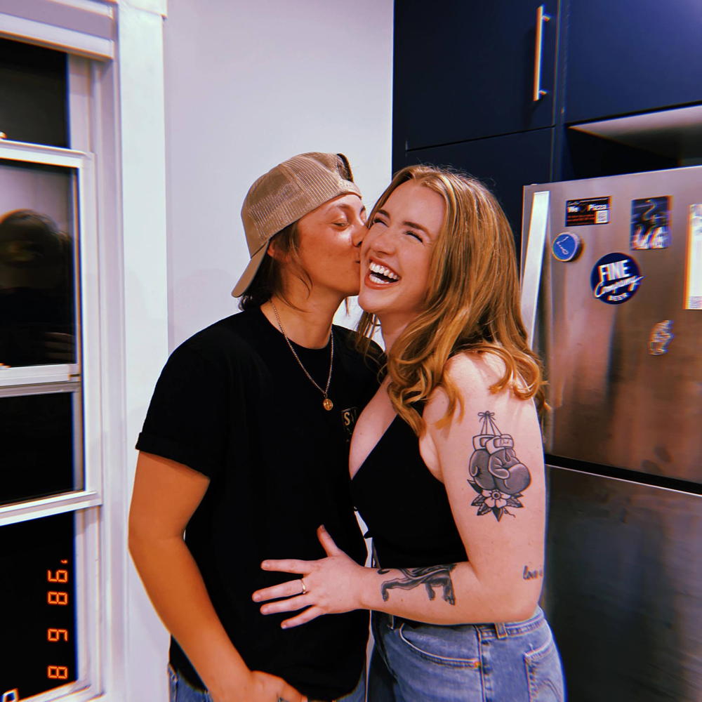 A woman in a halter top laughs as she's being kissed by another woman in a baseball cap.