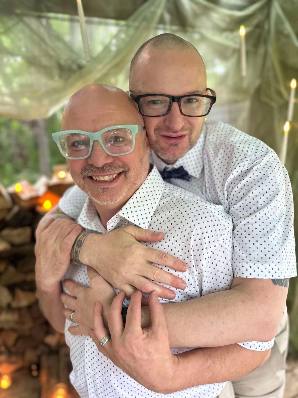 Jacob and steve embracing in an outdoor tent with candle type lights behind