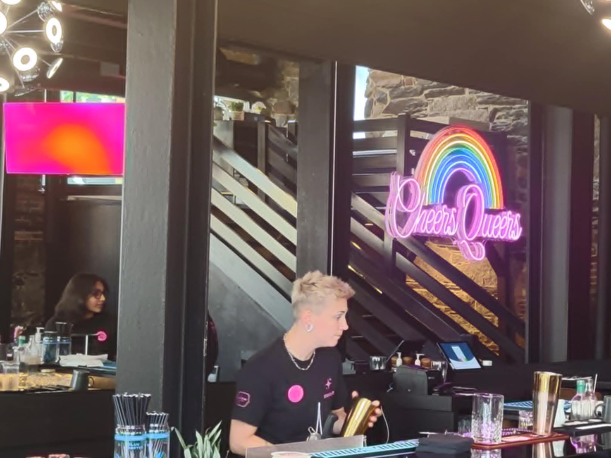 bartender at bar with "Cheers Queers" rainbow sign behind