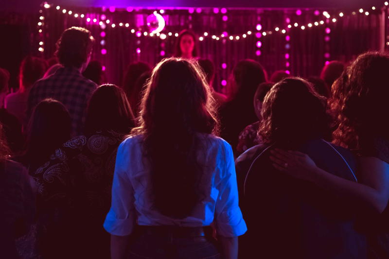 from the back of a nightclub. women watch a woman on stage surrounded by strings of white and purple lights