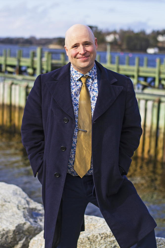 smiling man in jacket and tie and print shirt with a dock in the background