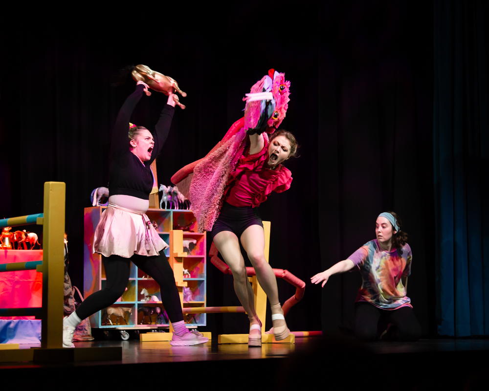three female characters on stage; the one with a satin apron on is brandishing a small cow